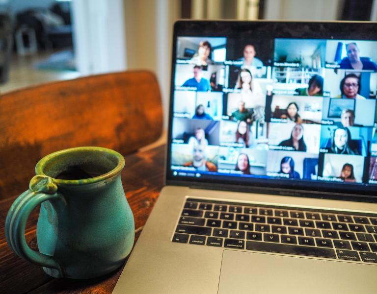 Coffe mug next to laptop with Zoom showing faces blurred out on a video meeting