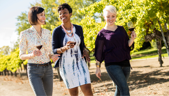 three women walking together in a vineyard holding wine glasses