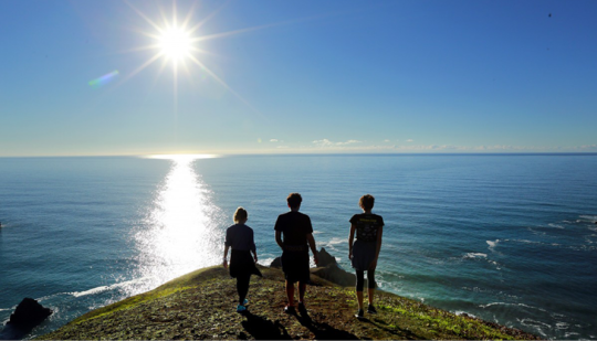 three people seen from behind walking towards a cliff edge above the sea