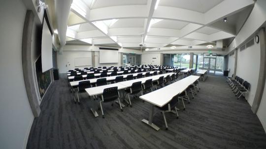 an interior classroom space with rows of white tables and black chairs with a white lattice ceiling and one wall full of windows